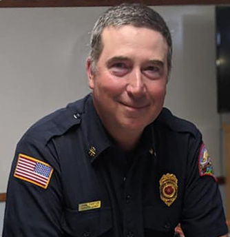 Fire Chief Jamie Painton smiling in a photo, wearing his dark blue uniform with patches and gold badge and nameplate.